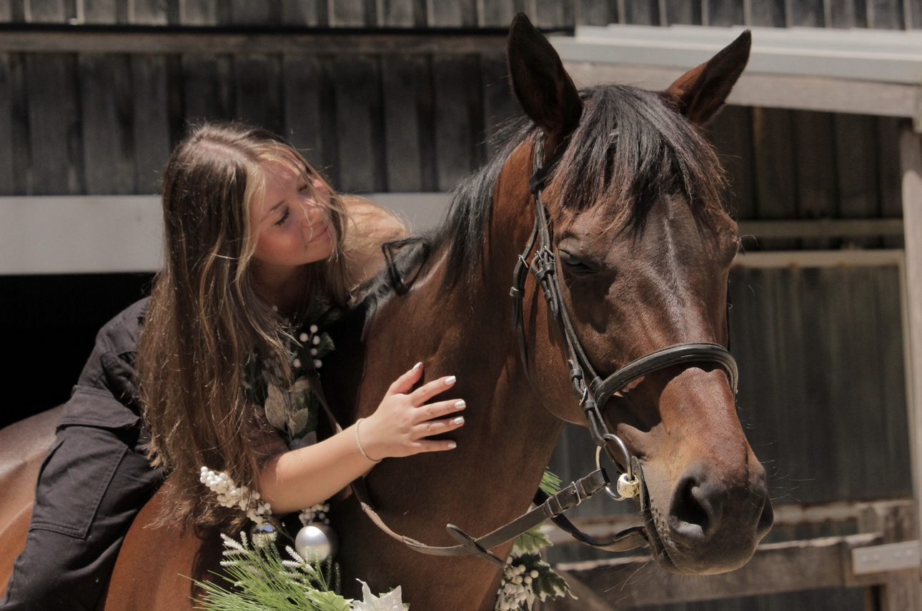 Ruby leaning over and patting her bay horse at the stables